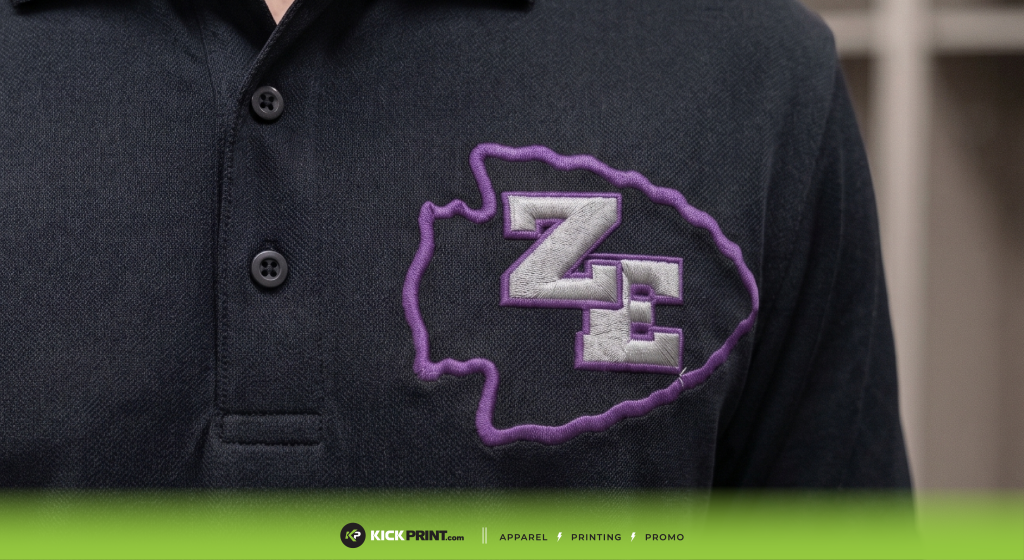 A close-up of a black polo shirt highlights detailed embroidery: a purple and white arrowhead outline with the letters ZE inside. The bottom edge features a green banner with the Kick Print logo and site navigation text.