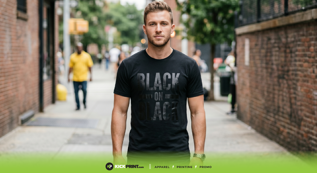 A man stands outdoors on a city sidewalk, wearing a black T-shirt with the words BLACK ON BLACK printed in bold Black Ink. The background shows blurred pedestrians, trees, and brick buildings. KickPrint.com branding is at the bottom.