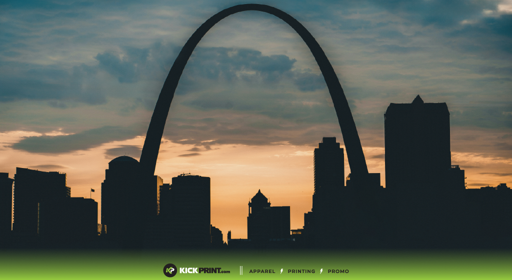 Silhouette of the St. Louis Gateway Arch and city skyline at sunset, with a dramatic sky and clouds in the background. A green banner at the bottom features the KickPrint logo, website, and highlights custom t-shirts for your company.