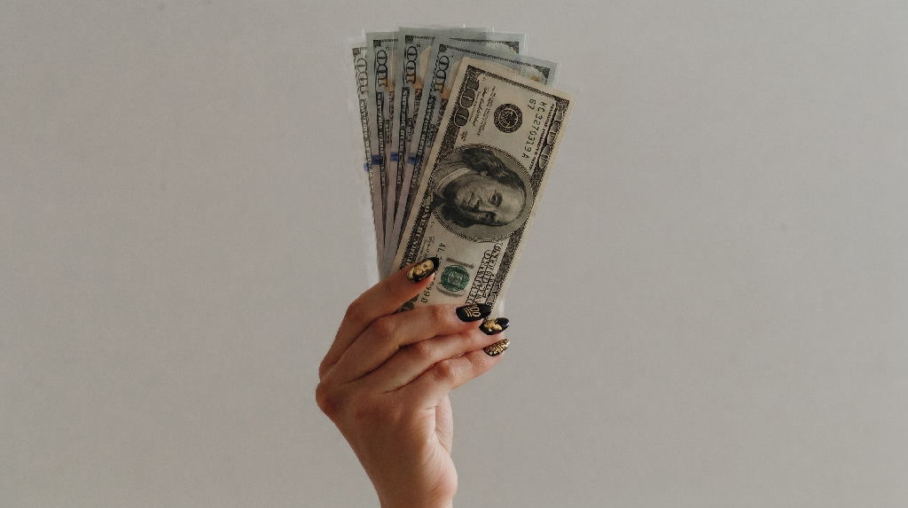 A hand with painted nails holds up a fan of U.S. hundred-dollar bills against a plain, light background, capturing the spirit of successful fundraising.