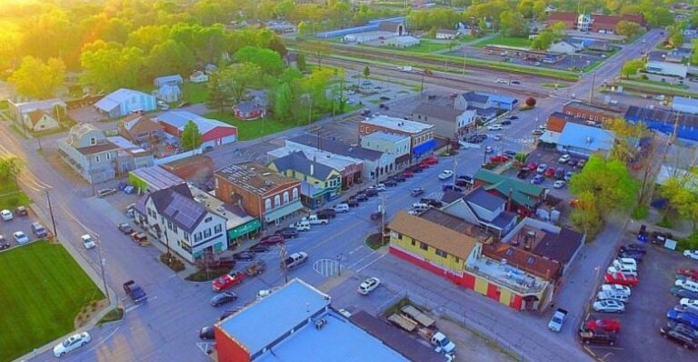 Aerial view of Eureka, a small town with colorful buildings, a main street lined with parked cars, green trees, and railroad tracks in the background on a sunny day.