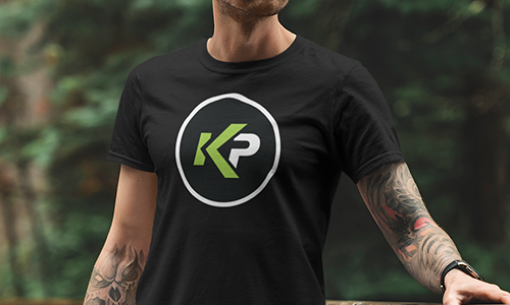 A person wearing a black t-shirt with a circular logo featuring the letters K in green and P in white, standing outdoors with trees in the background. The person has tattoos on their arm.
