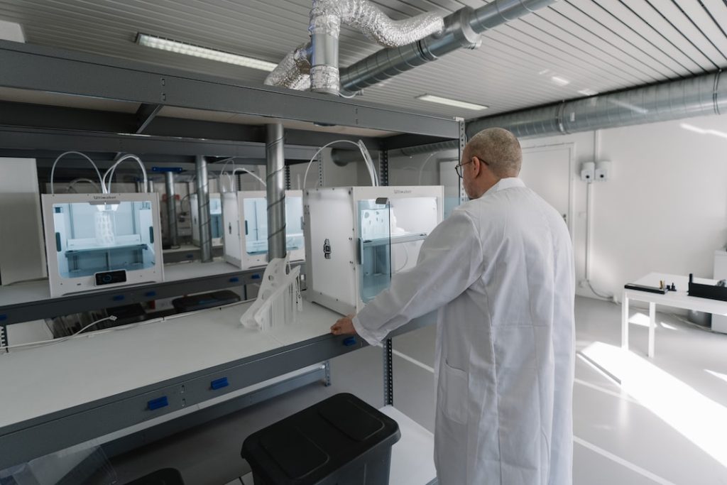 A person in a lab coat operates a white 3D printer in a modern laboratory with metal shelves, large windows, and visible air ducts.