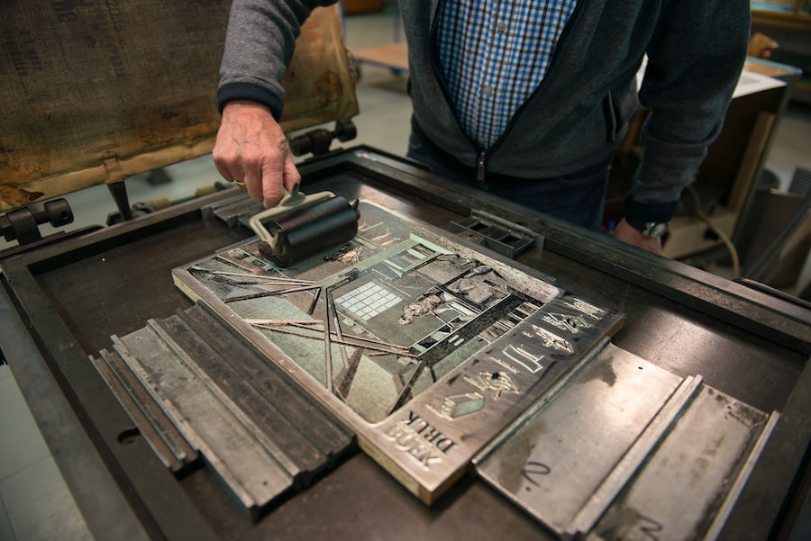 A person uses a roller to apply ink to a printing plate in a printmaking studio, preparing for the printing process. The plate features an intricate design and various letters.