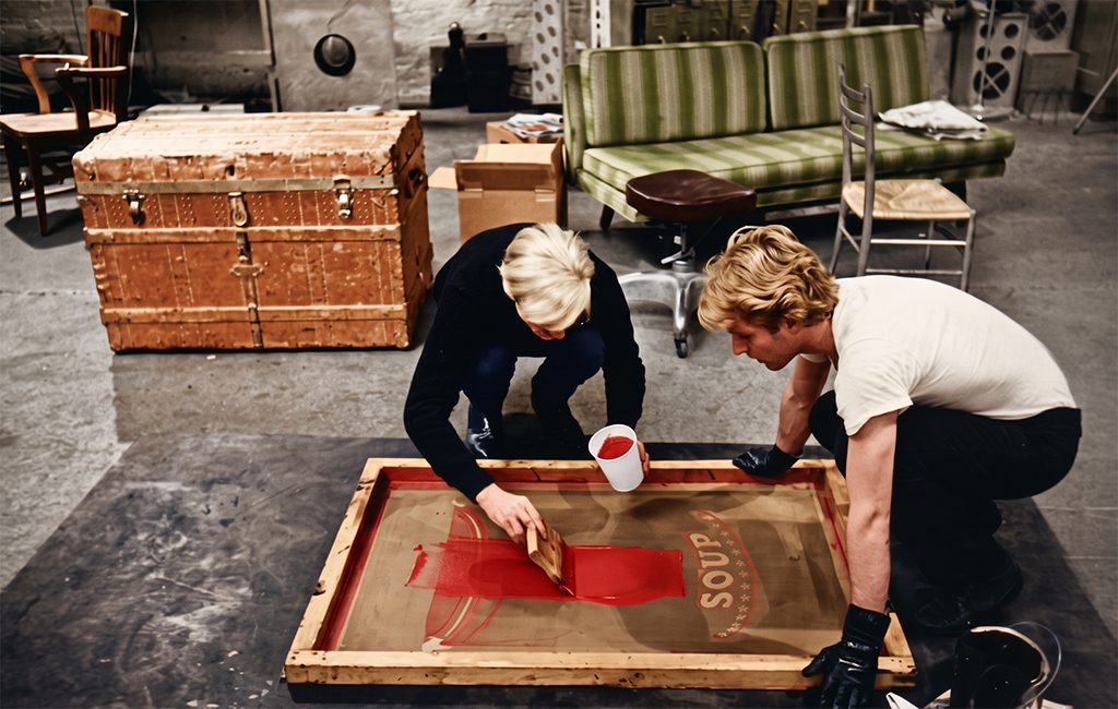 Two people collaborate on a screen printing project, spreading red ink over a large screen in a studio filled with vintage furniture and wooden crates, capturing the art and history of this creative process.