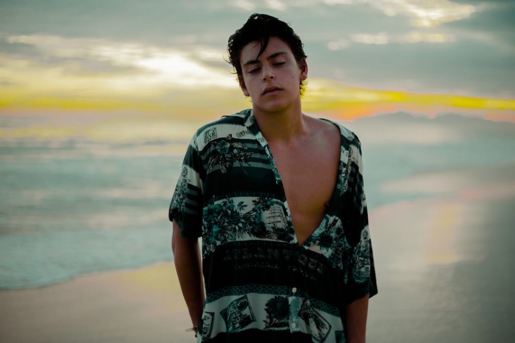 A person with short dark hair wearing an open patterned shirt stands on a beach at sunset, with waves and a cloudy sky in the background.