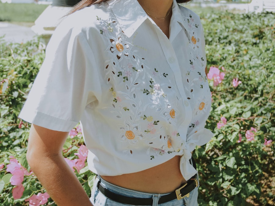 A person wearing a white, short-sleeved, collared, button-up shirt with floral embroidery and eyelet details, tied at the waist, standing in front of blooming pink flowers and greenery.