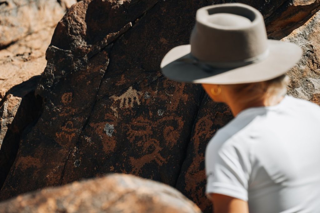A person in a wide-brimmed hat examines ancient petroglyphs carved into a brown, rocky surface under sunlight.