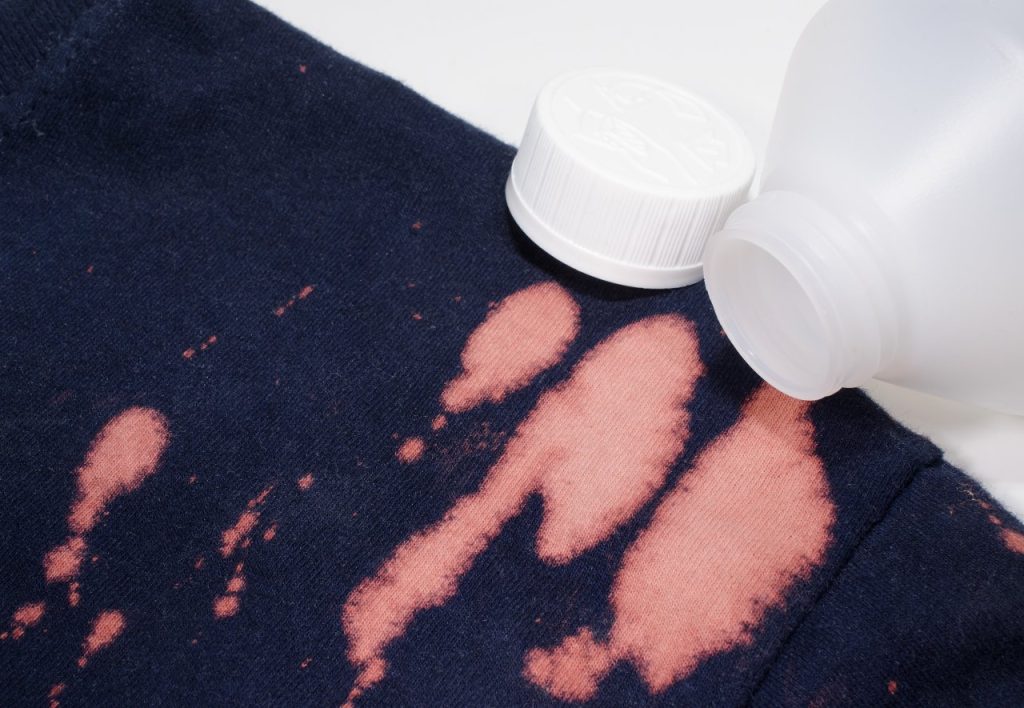 A navy blue fabric with large pinkish bleach stains is shown, alongside an open plastic bottle of bleach with its cap resting nearby—a reminder that shielding your shirts is crucial for safeguarding and cleansing amid pressure washing.
