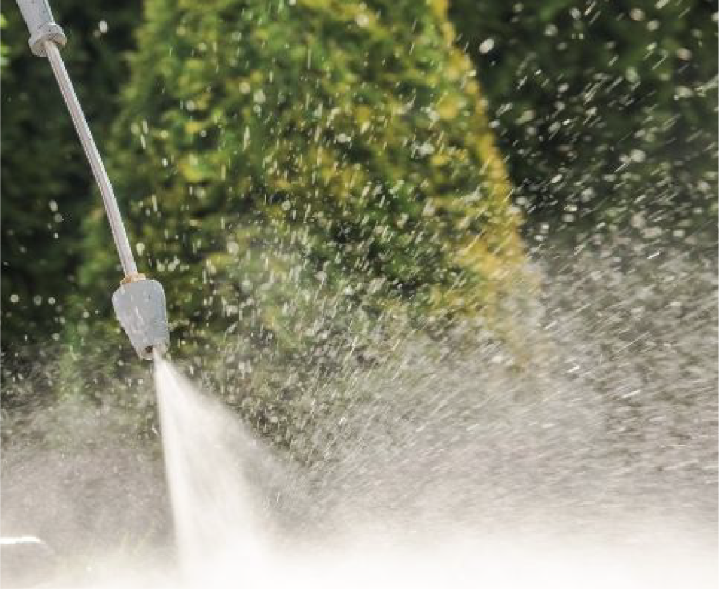 A close-up of a power washer spraying water, with shielding your shirts: safeguarding and cleansing amid pressure washing. Water mist and droplets are visible, while green trees blur in the background, emphasizing the force of the water jet.