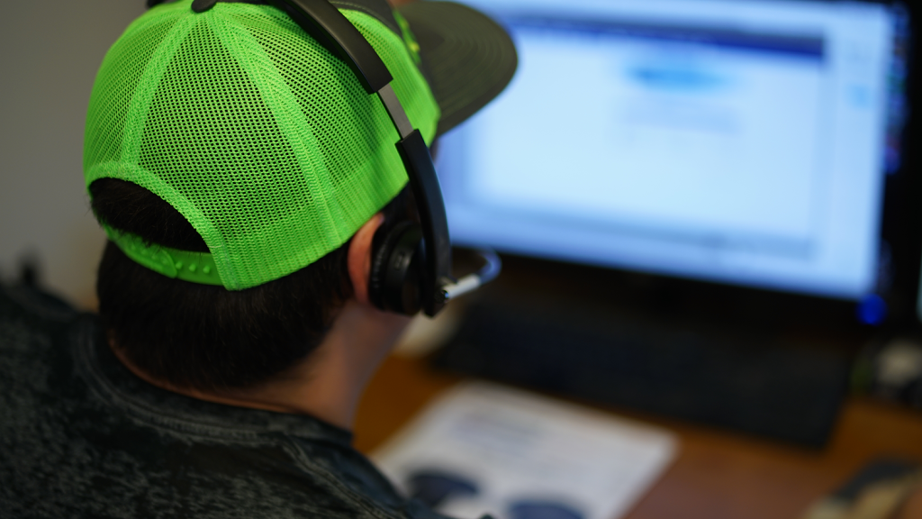 A person wearing a bright green baseball cap and headset sits at a desk, looking at a computer screen with blurred content. A paper with printed text is visible, hinting at Kick Print: Kirkwoods Premier Destination for Screen Printing Excellence!.