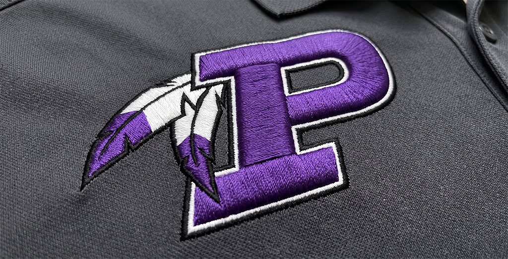 A close-up of a gray fabric with a large purple letter P outlined in white and black, featuring two white and purple feathers attached to the left side—showcasing detailed embroidery inspired by Kansas City style.