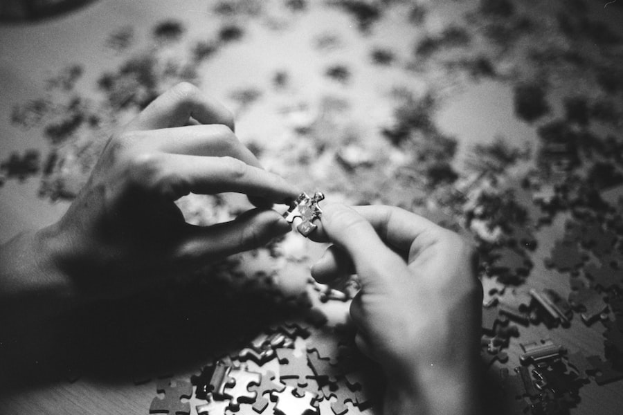 Close-up, black and white photo of two hands assembling a jigsaw puzzle on a table scattered with loose pieces. The focus is on the fingers holding a puzzle piece.