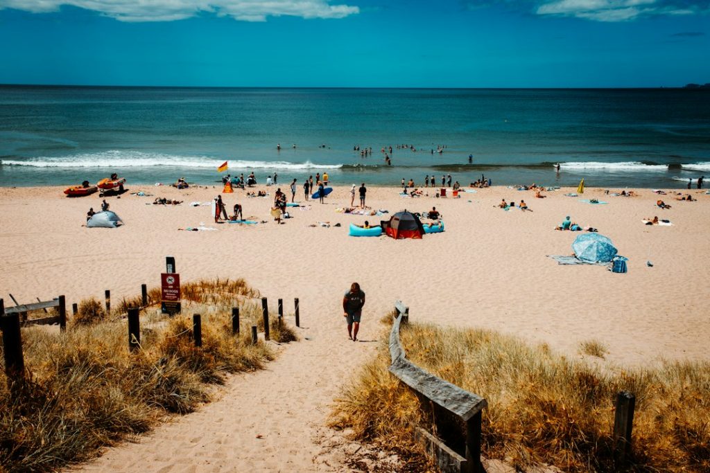 A person walks down a sandy path toward a busy beach, where people are sunbathing, swimming, and playing near the ocean under a blue sky. Some beach tents and umbrellas are set up on the sand.