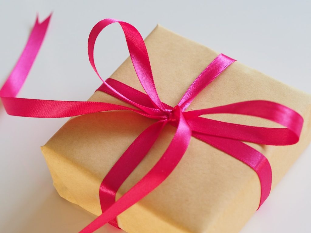A small gift box wrapped in brown paper and tied with a bright pink satin ribbon, forming a bow on top, set against a light background.