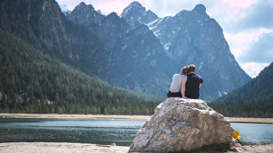 A couple sits together on a large rock by a lake, facing away from the camera and looking at tall, forested mountains under a partly cloudy sky.