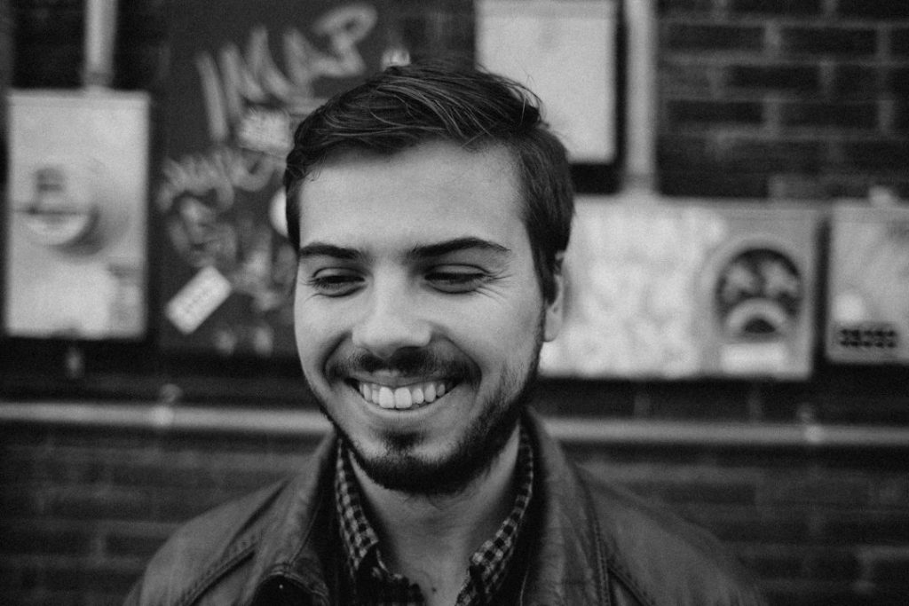 A young man with short hair and a beard smiles while looking down. He is wearing a checkered shirt and a jacket, standing in front of a brick wall with graffiti and electrical boxes. The image is in black and white.