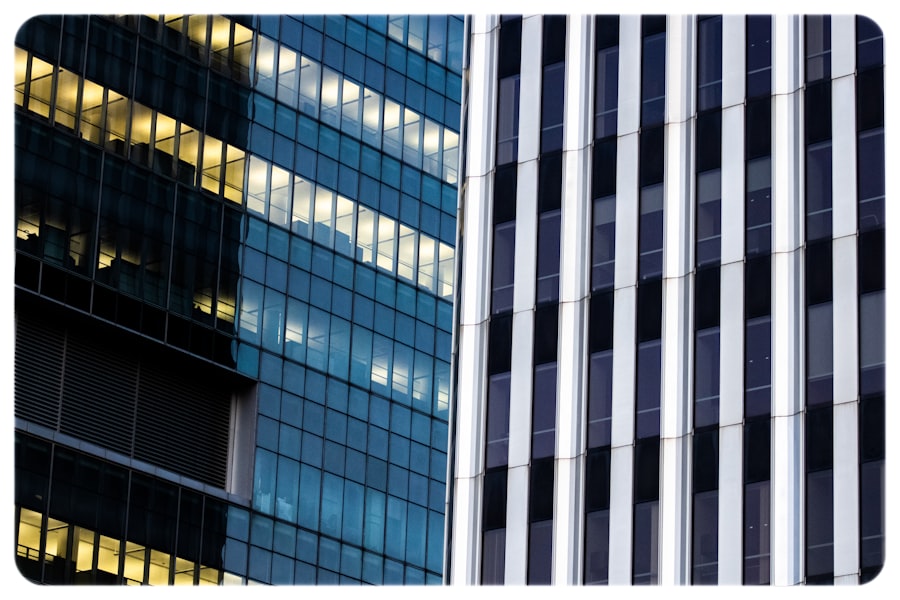 Close-up of two modern office buildings. The left building has blue-tinted glass windows with some lights on inside, while the right building features vertical white and dark gray stripes.