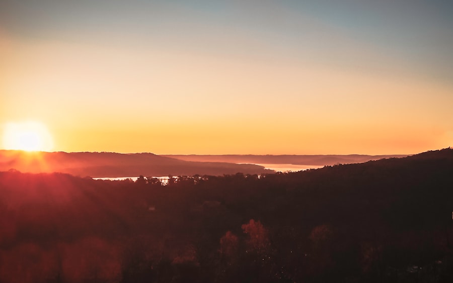 Sunrise over a landscape with dark, tree-covered hills in the foreground, a river or lake reflecting light in the middle, and a clear sky with warm orange and yellow hues on the horizon.