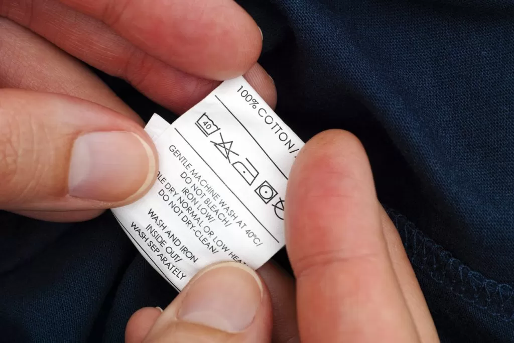 Close-up of hands holding a clothing care label on dark fabric, featuring washing and ironing instructions—Mastering T-Shirt Care: Essential Dos and Donts for Maintaining Your Wardrobe Staples like warnings to avoid fire, bleach, or dry cleaning.