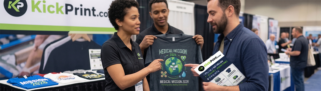 Three people at a KickPrint.com screen printing company booth at a convention; one woman and one man display a “Medical Mission 2025” t-shirt to another man holding flyers, with shirts displayed on the table.