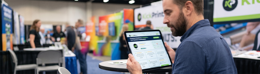 A man sits at a table holding a tablet and looking at its screen during a business expo, with colorful booths—including a prominent screen printing company— and people in the busy background.