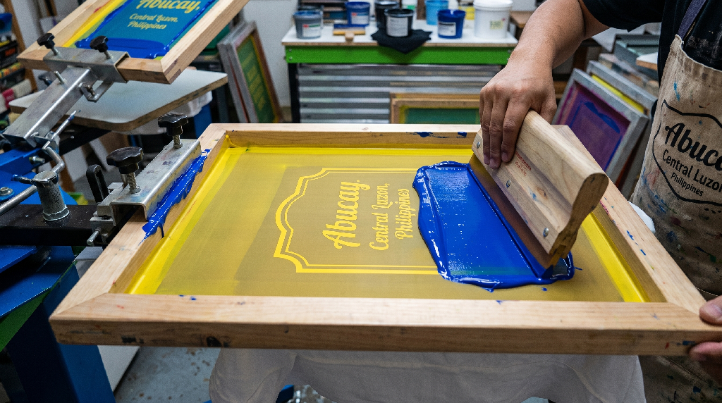 A person screen prints a yellow Abucay, Central Luzon, Philippines design onto fabric, spreading blue ink over a stencil with a squeegee in a workshop close to me filled with printing materials.
