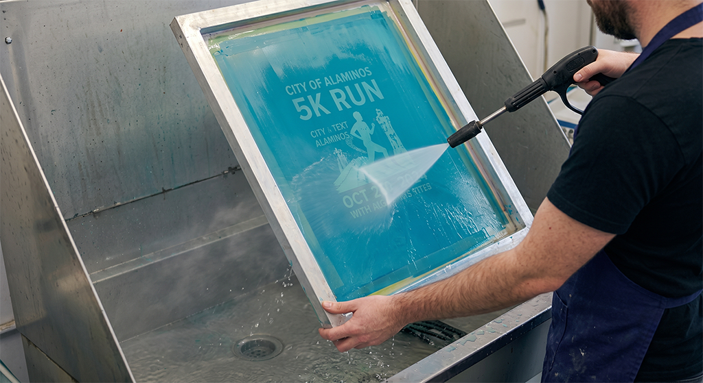 A person uses a spray hose to clean a screen printing frame featuring a City of Alaminos 5K Run design at a workstation sink, showcasing the quality of local screen printing services.