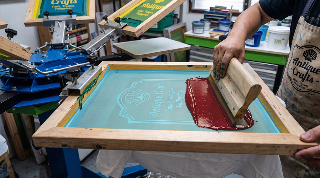 A person is screen printing a t-shirt by spreading red ink over a stencil with a squeegee. The stencil reads “Antique Crafts.” Various printing tools and materials are visible, much like those found in nearby screen printing shops.
