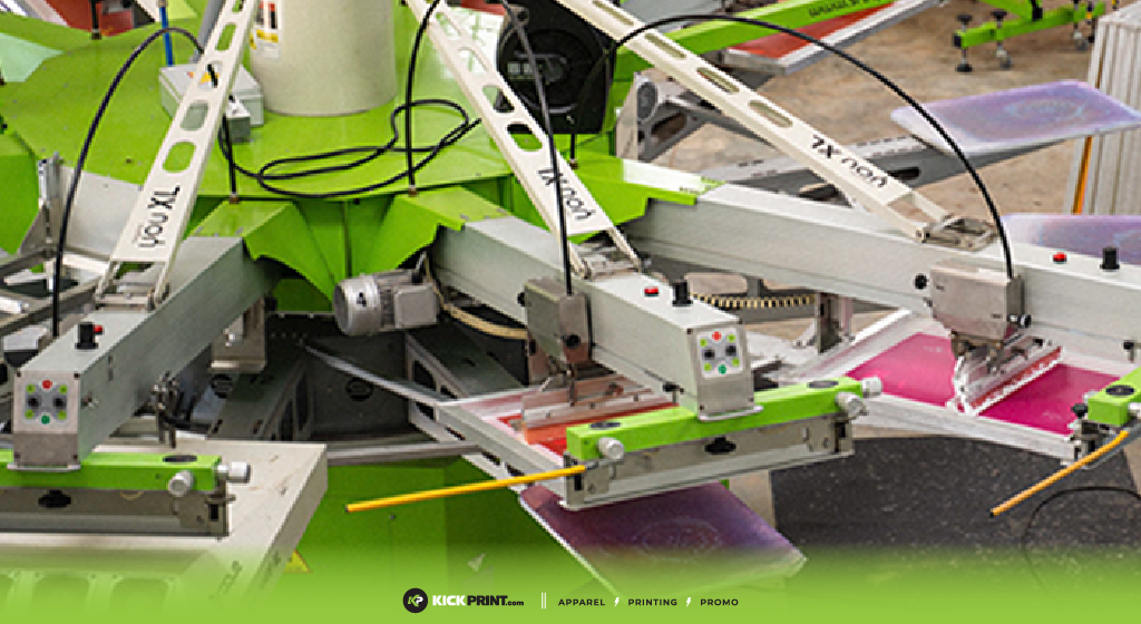 A close-up of a screen printing machine with multiple arms and screens, printing colorful designs onto fabric. The background shows part of a Missouri workshop. The Kick Print logo and website appear at the bottom, highlighting top Screen Printing Companies.