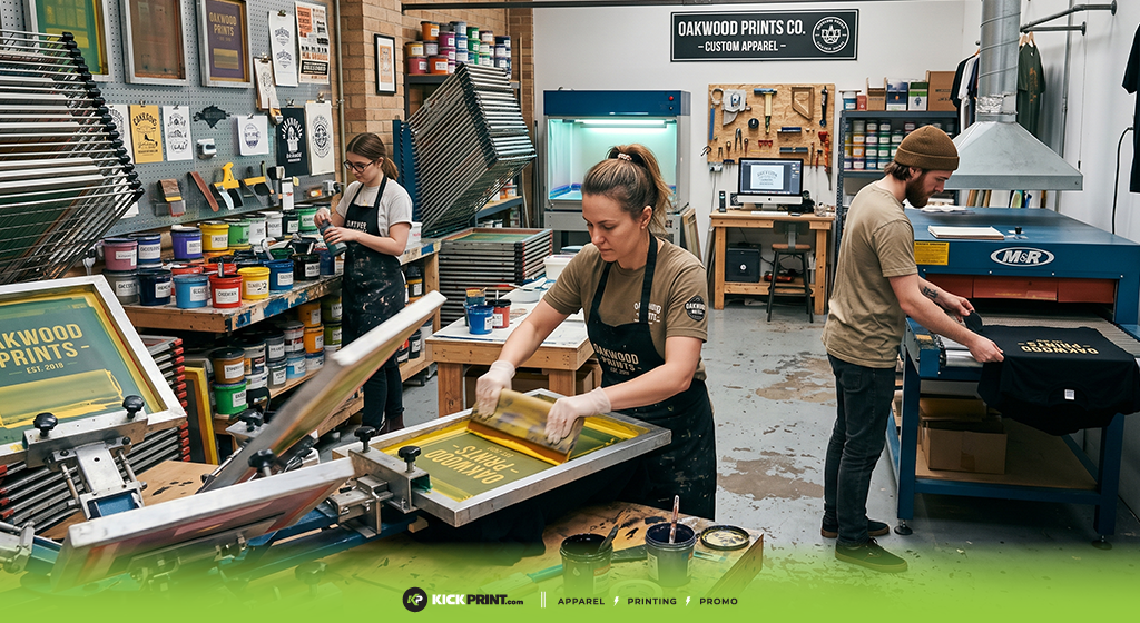 Three people work in a screen printing shop, applying ink to shirts and handling equipment. Colorful ink containers line shelves, and finished shirts for custom wardrobes are stacked nearby. The space is organized and filled with printing tools.
