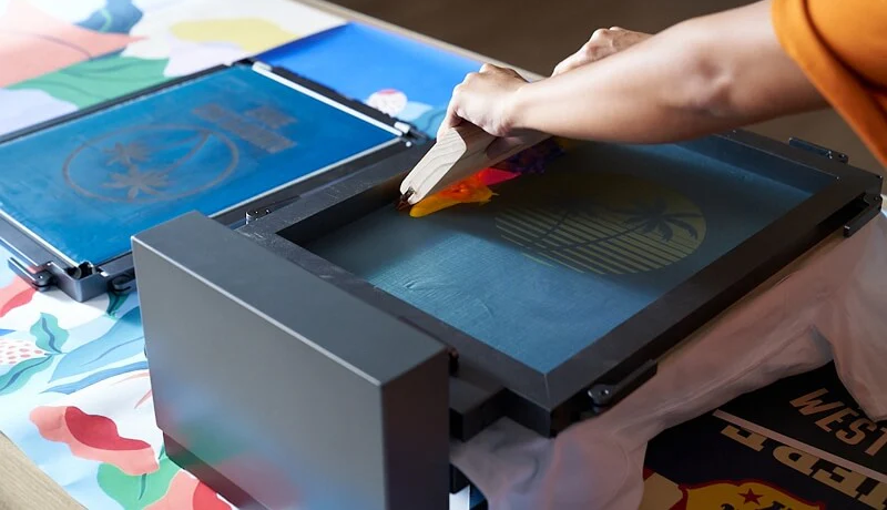 A person uses a squeegee to spread ink across a screen for professional screen printing, transferring a design onto a white fabric positioned under the screen frame.
