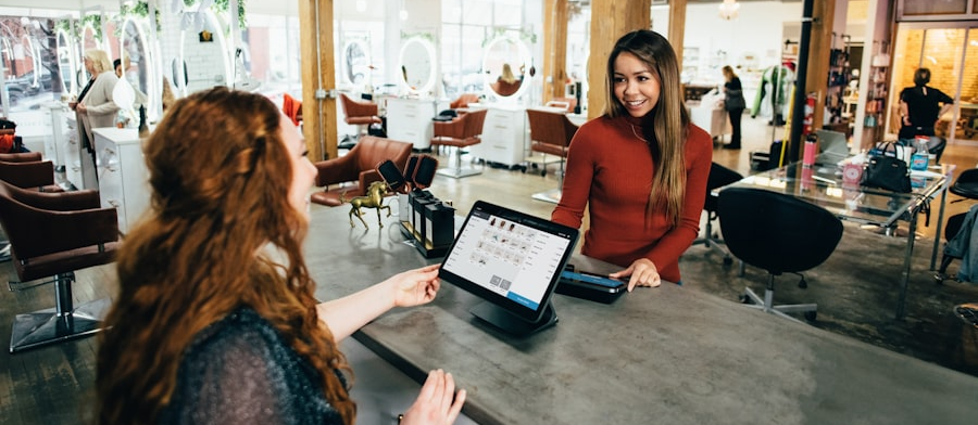Two women interact at a salon counter; one stands behind the counter, smiling, while the other gestures towards a touchscreen register. Salon chairs and mirrors are in the background of this stylish beauty spot near me.