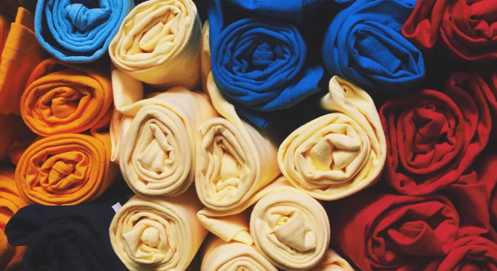 Rolled fabrics in bright yellow, blue, orange, cream, and red are arranged in a grid pattern, creating a vibrant display from above—perfect inspiration for affordable screen printing and quality prints.
