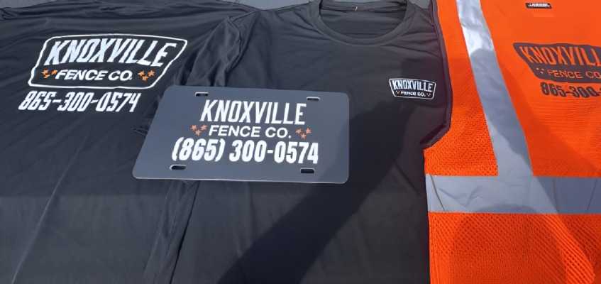 Black T-shirts, a license plate, and an orange safety vest—each branded with Knoxville Fence Co. and the phone number (865) 300-0574—are neatly laid out on a flat surface.