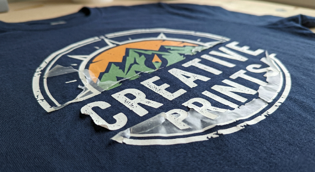 A navy blue t-shirt with a colorful mountain and sun logo and the words Creative Prints, partially covered by clear transfer tape, ready for heat pressing with DTF transfers.