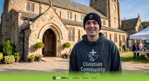 A smiling young man in church merch—a Christian Community Church hoodie and beanie—stands in front of a stone church building. The scene is bright, with potted plants near the entrance and a tent visible to the right.