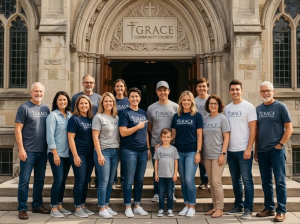 A group of people wearing matching church t-shirts stand smiling together on the steps outside a stone church building with an arched entrance and a sign reading Grace Community Church above the door.
