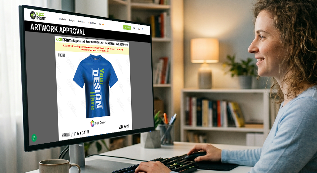 A woman sits at a desk, smiling at a computer screen displaying an artwork approval page from a T-Shirt Printing Company for a blue custom T-shirt design that says YOU DESIGN HERE.