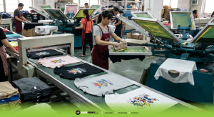Workers in a T-shirt printing company print colorful designs on shirts using large machines and conveyor belts, with several shirts in progress and printed ones laid out to dry for the fastest T-shirt printing results.